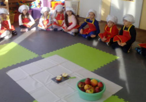 Dzieci w fartuszkach i czapkach kucharskich siedzą w kole. W środku koła ustawiona miska z jabłkami i jabłko rozkrojone.